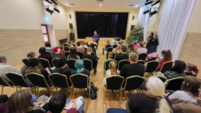 Tautas nama zālē Ķeguma audzēkņi sēž uz krēsliem un pedagogs runā priekšā uz skatuves