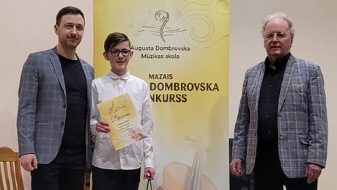 zēns baltā kreklā, turot diplomu, ar 2 vīriešiem nostājušies pie dzeltenīga banera