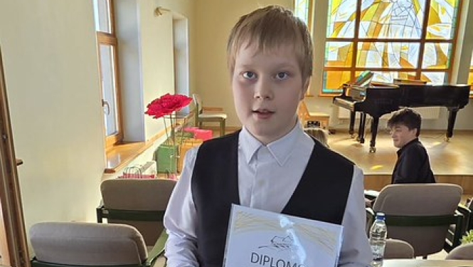 zēns ar gaišiem, īsiem matiem, svētku apģērbā tur diplomu