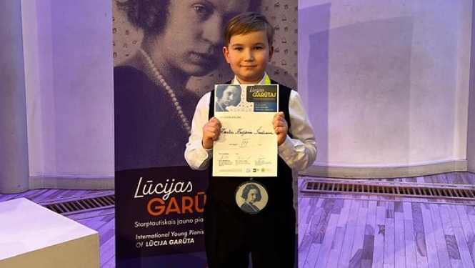 zēns svētku drēbēs ar medaļu un diplomu nostājies pie Lūcijas Garūtas Starptautiskā jauno pianistu konkursa banera