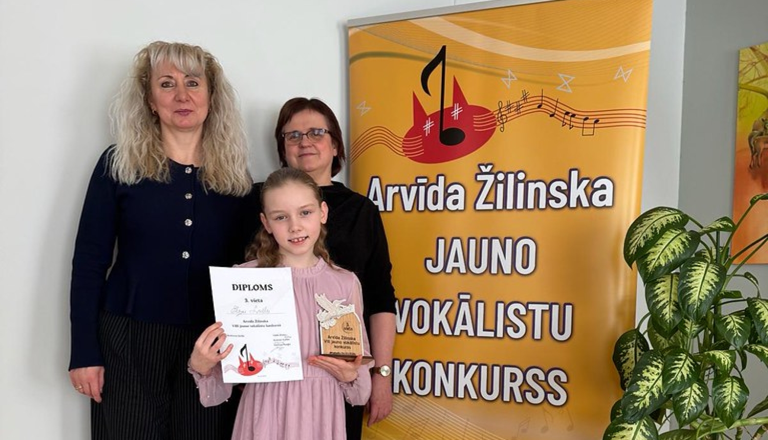 meitene rozā kleitā kopā ar divām sievietēm nostājusies pie banera un tur rokās diplomu