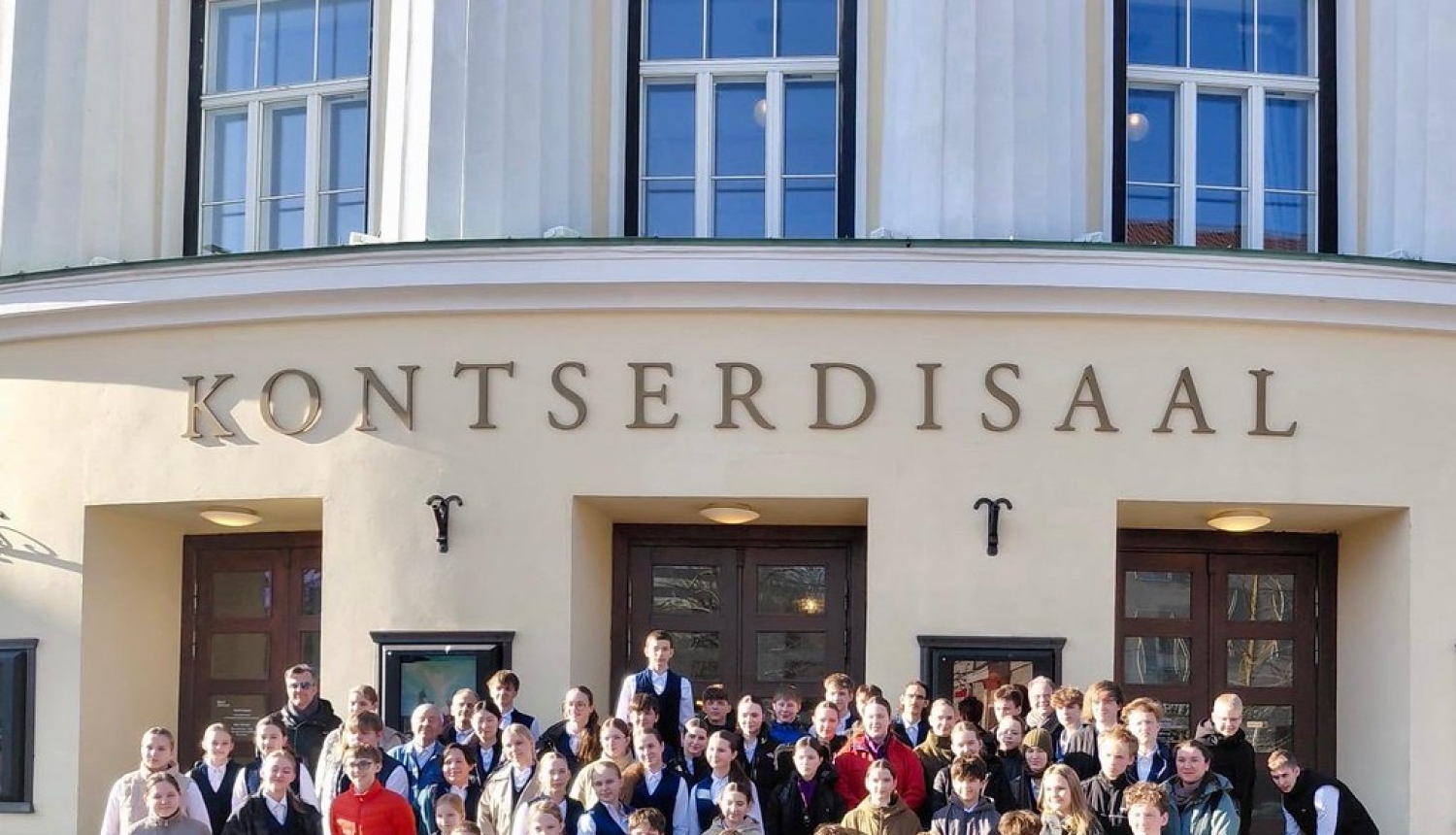 Pūtēju orķestris pozē priekšā koncertzāles uzrakstam Kontserdisaa