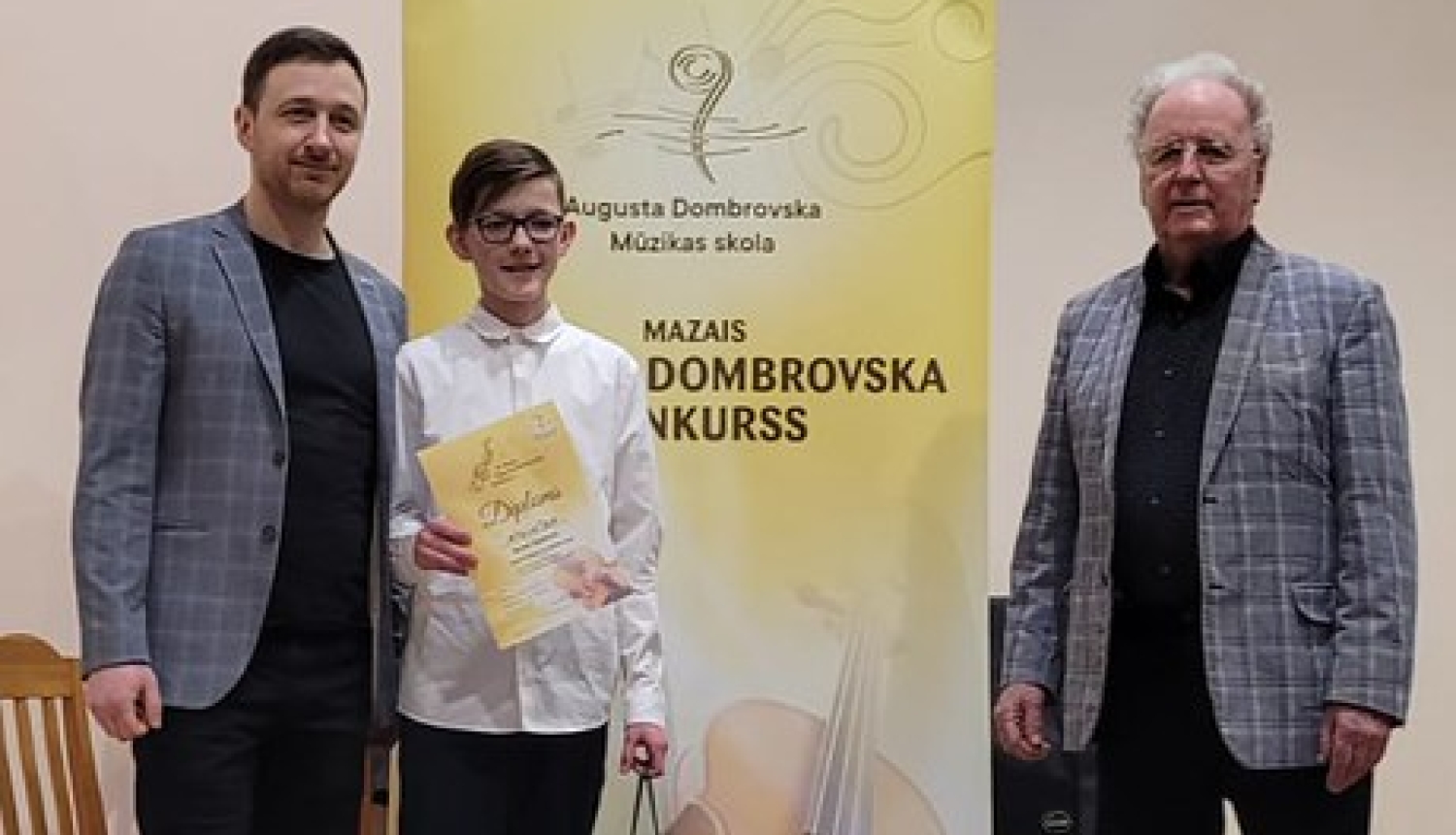 zēns baltā kreklā, turot diplomu, ar 2 vīriešiem nostājušies pie dzeltenīga banera