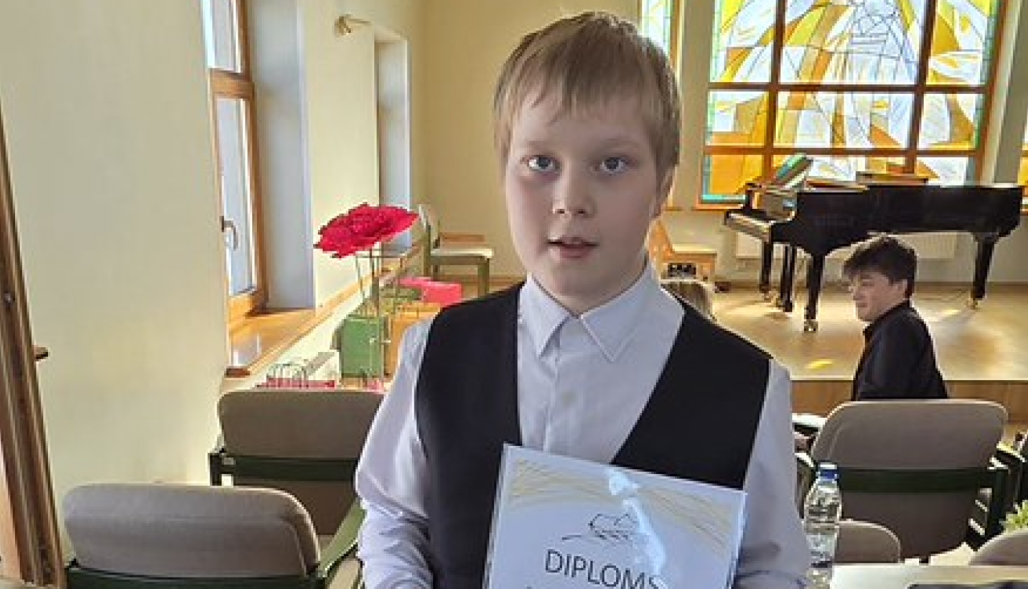 zēns ar gaišiem, īsiem matiem, svētku apģērbā tur diplomu