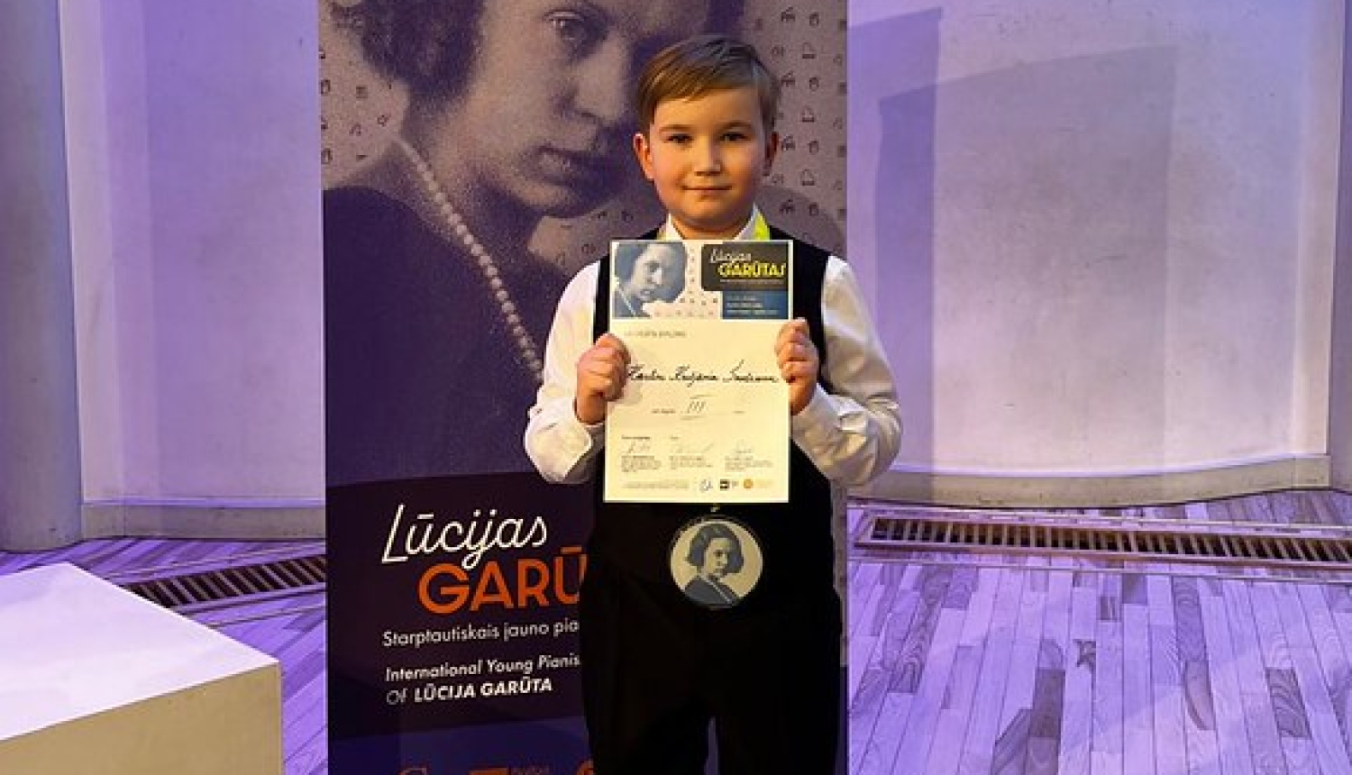 zēns svētku drēbēs ar medaļu un diplomu nostājies pie Lūcijas Garūtas Starptautiskā jauno pianistu konkursa banera