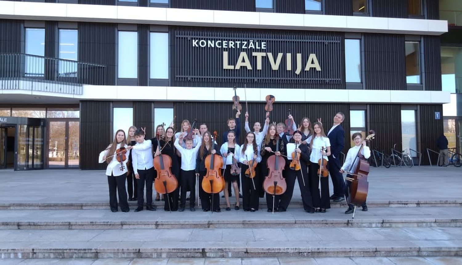 Stīgu kamerorķestris ar diriģentu nostājušies priekšā koncertzālei ,,Latvija’’