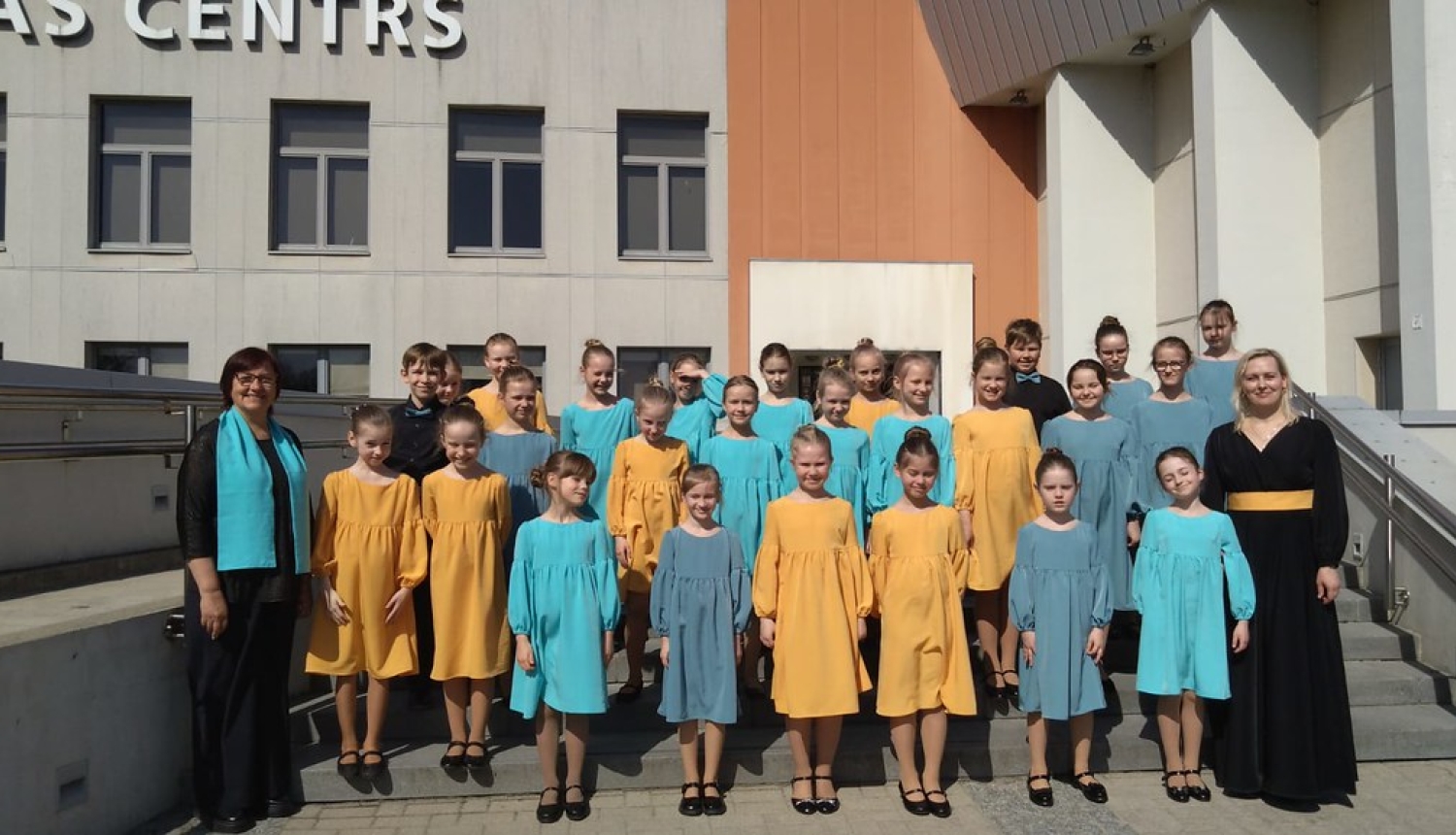 bērnu koris ar divām skolotājām saulainā dienā nostājušies uz kāpnēm